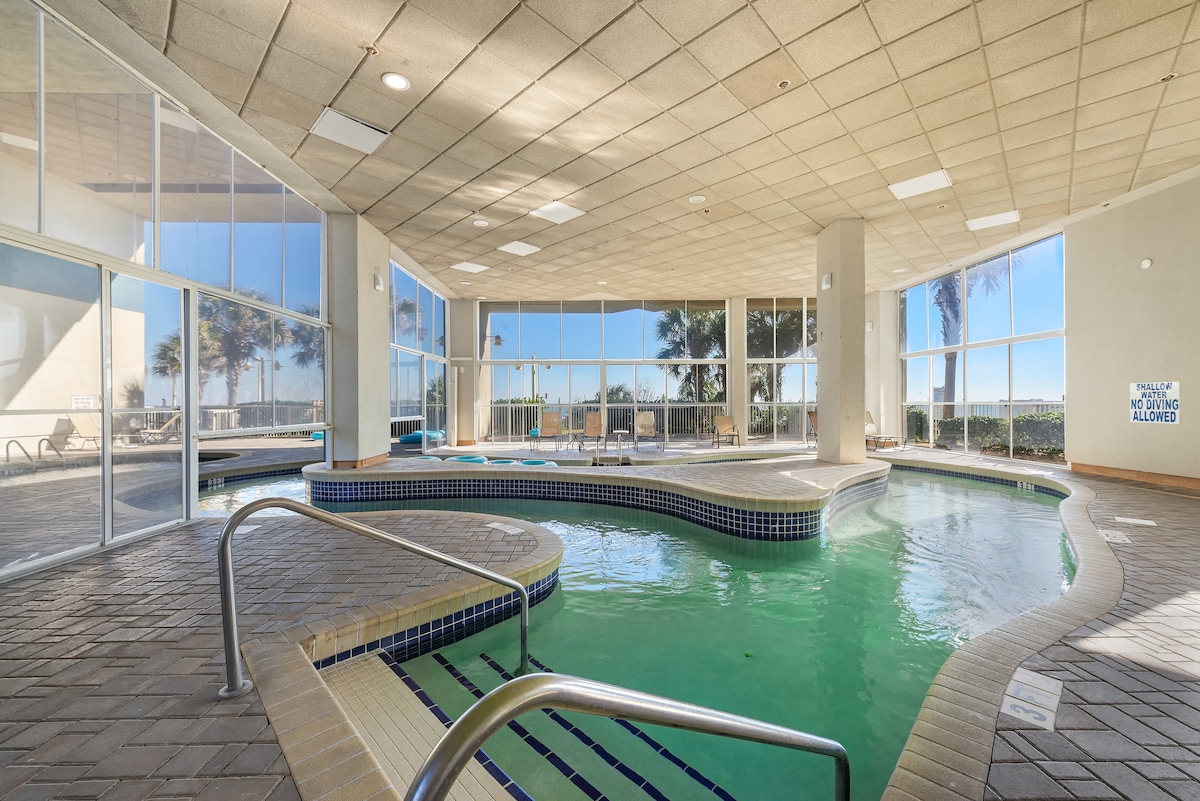 An indoor pool area is presented, featuring a winding structure with varying depths and a tiled edge. Large windows allow ample natural light, offering views of palm trees outside. Comfortable seating is visible in the background, creating a relaxing atmosphere.