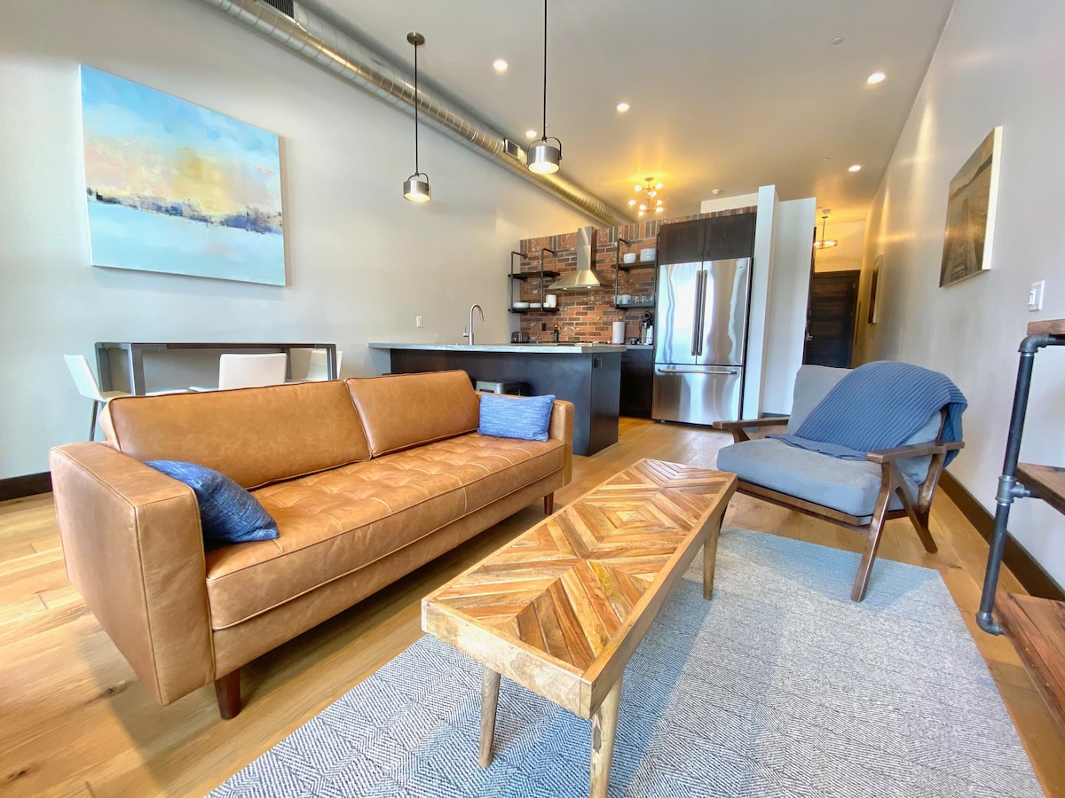 The open living space showcases a leather sofa accompanied by decorative pillows, paired with a wooden coffee table featuring a chevron design. A kitchenette with stainless steel appliances is visible in the background, complemented by modern lighting fixtures above. A chair with blue fabric adds a touch of color.