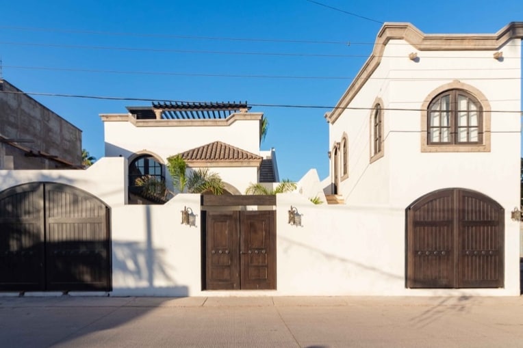 A charming exterior view of a white-walled property features distinctive dark wooden gates and arched windows. The roof terrace is visible, surrounded by greenery, showcasing a vibrant energy. Clear skies provide a bright backdrop, enhancing the design of the structure.