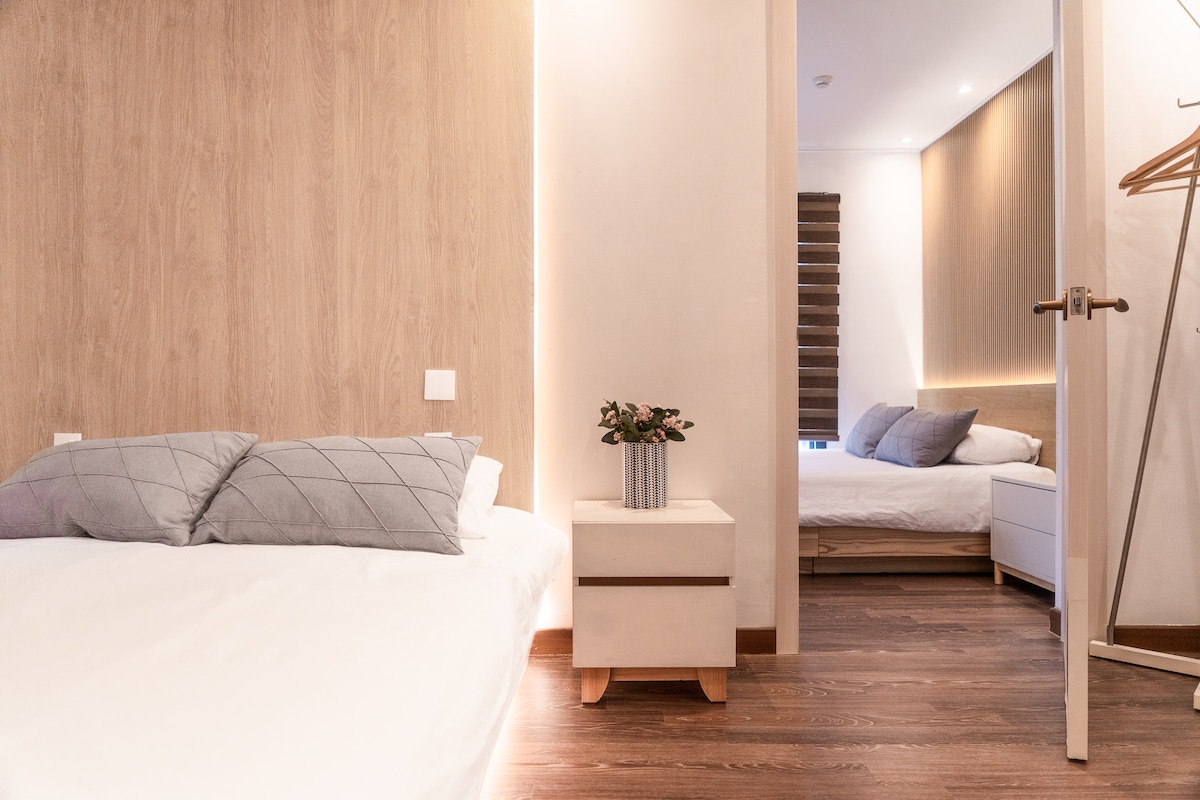 The image features a bright and inviting bedroom with light wood accents. A spacious bed is positioned to the left, complemented by a small bedside table with a vase. A second bed is visible in the adjacent room, providing additional sleeping space.