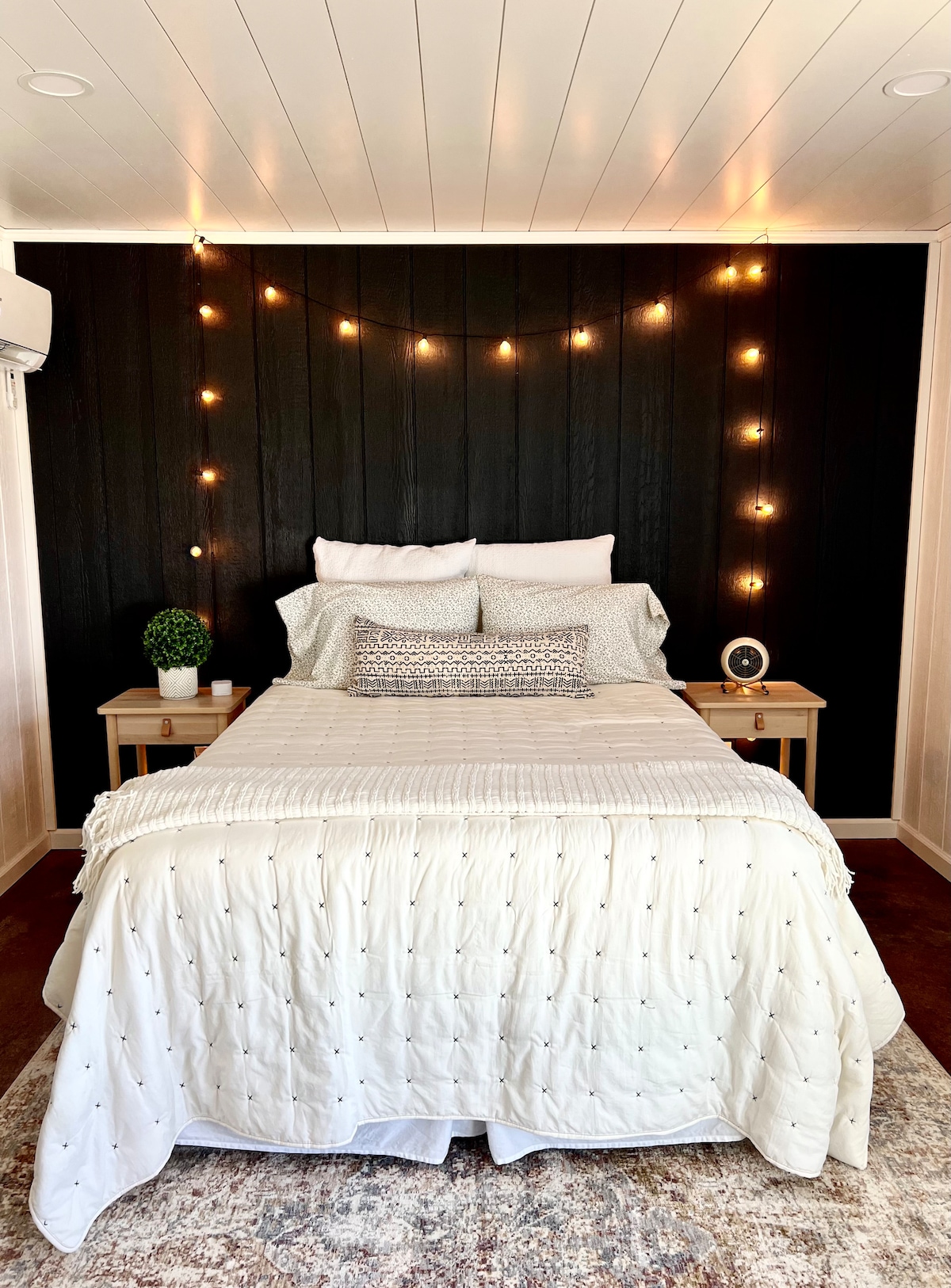 A cozy sleeping area features a spacious bed dressed in a light quilt, flanked by two bedside tables. Soft lighting from string lights enhances the dark accent wall, while a decorative plant and clock add subtle touches. A plush rug grounds the room.