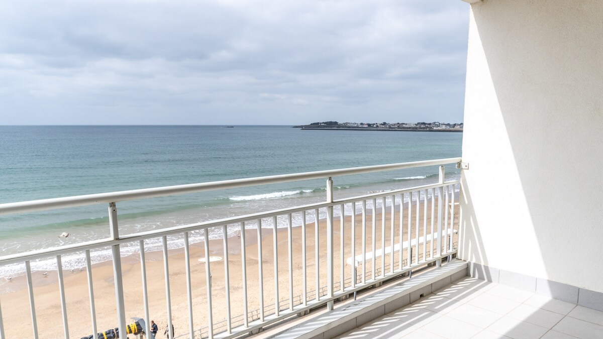 A spacious balcony overlooks the sandy beach and calm waters, extending towards the horizon. The railings provide a clear view of the shoreline and gentle waves, with a distant shoreline visible in the background.