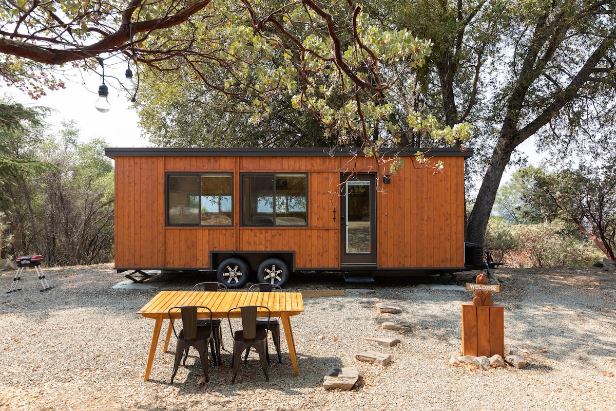 The tiny house, featuring a warm wooden exterior, is positioned on a gravel surface surrounded by trees. A wooden dining table with chairs is located in front, alongside a BBQ grill and a welcoming sign. Natural light fills the windows, providing a bright and inviting view.
