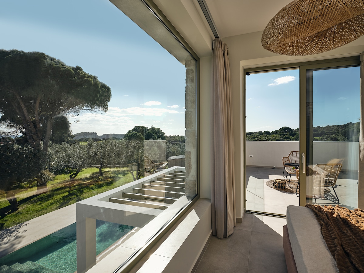 A sunlit corner of the villa is depicted, featuring large glass windows opening to a serene outdoor view. A cozy seating area includes soft textiles, while a woven light fixture hangs overhead. The outdoor landscape showcases lush greenery and the shimmering private pool.