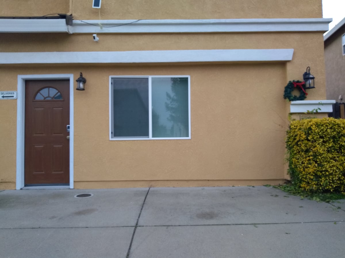 An exterior view of a private studio entrance is shown, featuring a wooden door with a small window above it. A large window beside the door allows natural light in, while lush greenery decorates the front landscape, complementing the beige exterior walls.