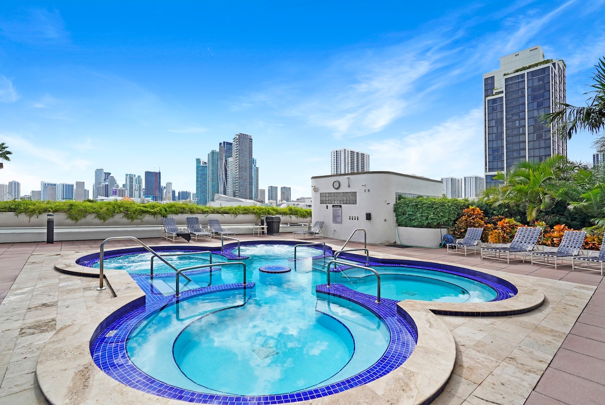 A rooftop pool area features two hot tubs and a spacious swimming pool, surrounded by lounge chairs. Lush greenery lines the perimeter, while the backdrop showcases the impressive skyline of downtown Miami under a clear blue sky.