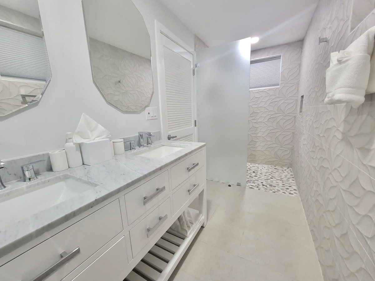 A modern bathroom features dual sinks with a marble countertop and sleek cabinetry. A spacious shower area displays a textured wall design and a pebble floor, while soft lighting enhances the overall ambiance. Fresh towels are neatly arranged on a rack.
