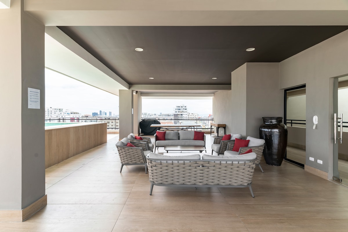 A spacious outdoor terrace is furnished with comfortable seating, accentuated by decorative cushions. A large vase adds a touch of elegance. The area offers expansive views of the city skyline, with light pouring in from ample windows, ensuring a bright and airy atmosphere.