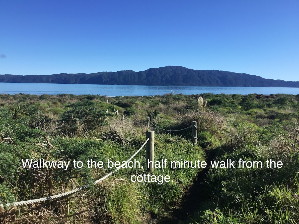 A winding walkway is visible leading through grassy terrain towards the beach. The shoreline and distant hills are seen in the background, creating a serene natural setting. The image conveys the accessibility of the beach, located a short stroll from the cottage.