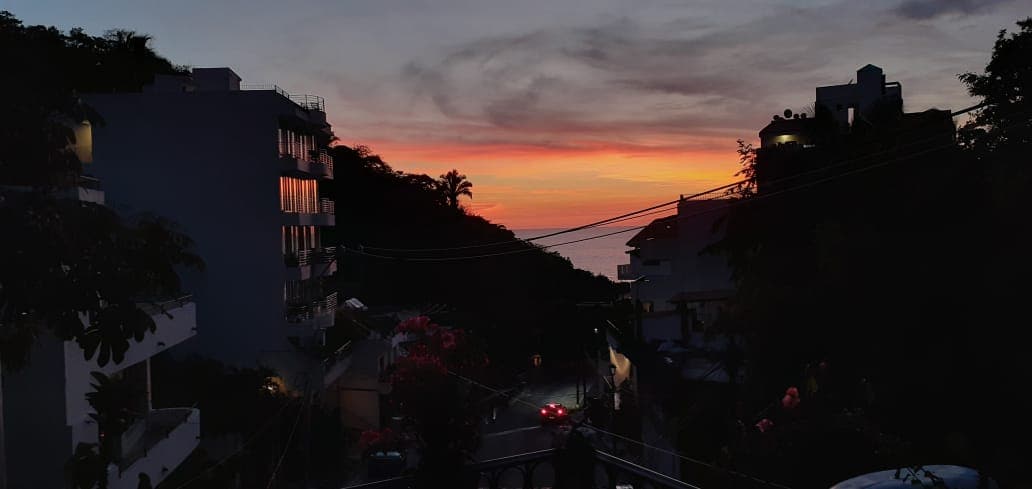 A serene evening scene captures the sunset over the horizon, with vibrant colors reflecting off buildings. Street lights create a soft glow along the road, while silhouettes of palm trees are visible against the colorful sky, providing a tranquil atmosphere.