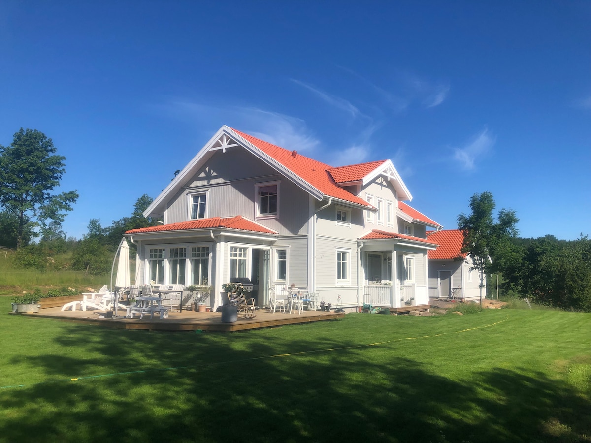 A spacious two-story house is shown with a light-colored exterior and a red-tiled roof. The surrounding area features a well-maintained lawn and a deck with seating arrangements. Trees and greenery create a natural backdrop under a clear blue sky.