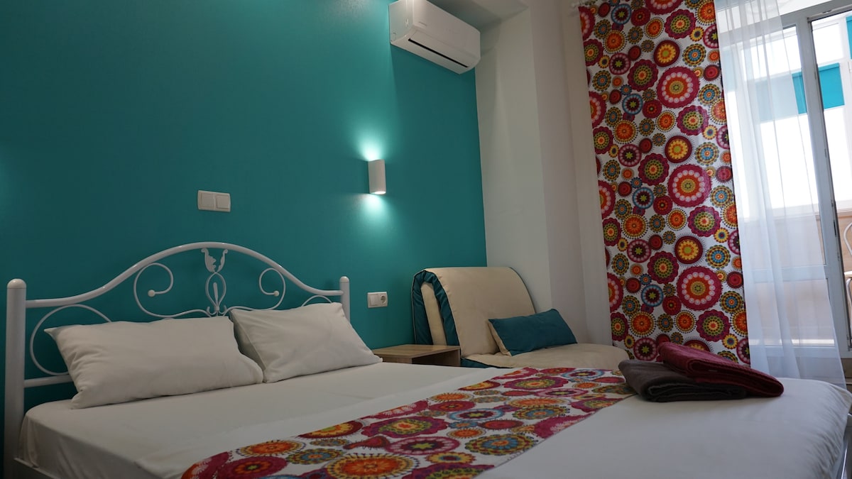 A smart apartment features a spacious bed adorned with a colorful patterned runner. A relaxing chair is positioned nearby, with a plush cushion adding comfort. Vibrant turquoise walls frame the area, while natural light filters through sheer curtains, ensuring a bright atmosphere.