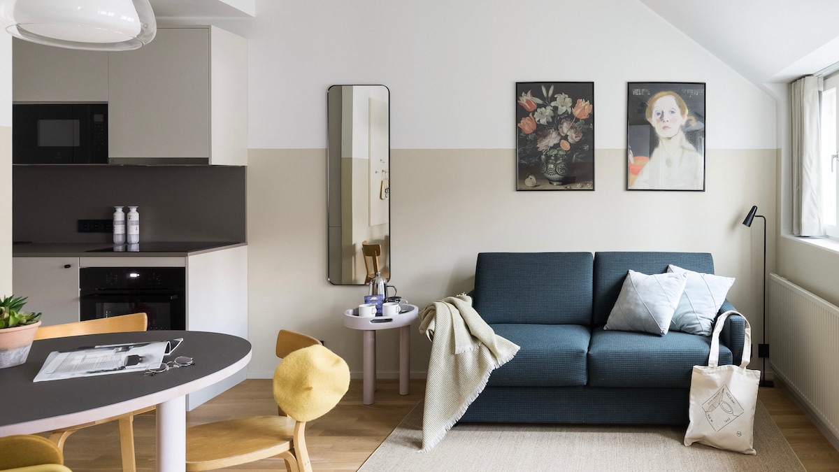A modern living area features a comfortable sofa complemented by a light throw and decorative pillows. A circular coffee table is placed beside it, while a dining table is visible in the foreground. Natural light enters through large windows, illuminating the elegant wall art.