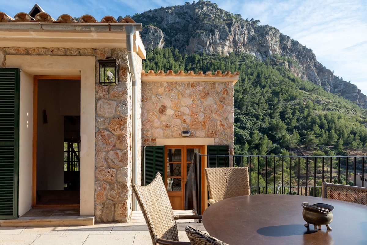 A stone exterior features rustic charm, complemented by green shutters and a tiled roof. A spacious terrace area is furnished with a round table and woven chairs, inviting relaxation. The backdrop showcases a mountainous landscape, lush with trees under a clear blue sky.