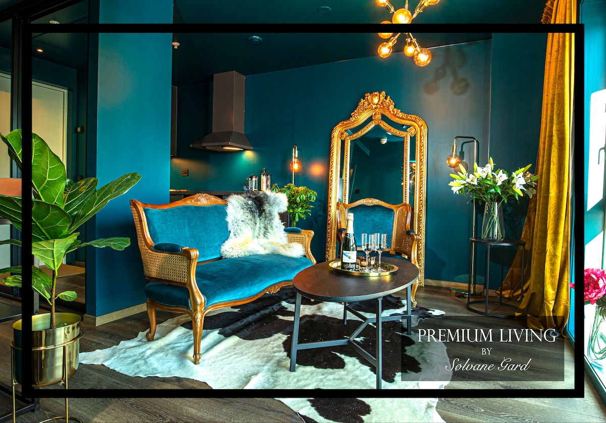 A stylish seating area features a plush blue sofa and an ornate gold-framed mirror. The floor is adorned with a patterned area rug, while ambient lighting highlights the deep blue walls, creating a cozy and inviting space.