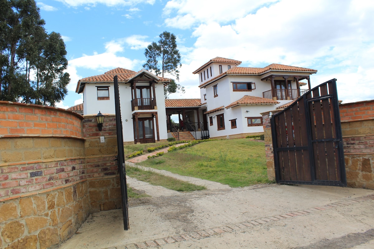 An elegant two-house setup is visible, showcasing a blend of modern and traditional architecture. The property is enclosed by a rustic stone wall and features a charming wooden gate. Lush greenery surrounds the houses, contributing to a peaceful rural setting under a partly cloudy sky.