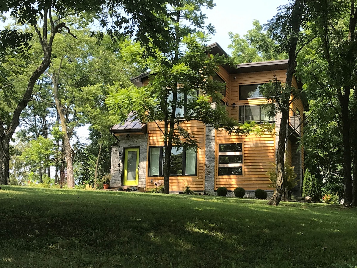 The two-story retreat is framed by lush greenery, showcasing a mix of wood and stone exterior. Large windows provide ample natural light, while a modern entryway is highlighted in a vibrant hue. The surrounding yard features well-maintained grass and mature trees.