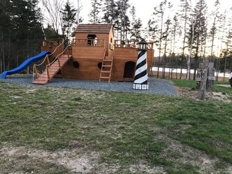 A wooden children's playscape resembling a pirate ship stands in a grassy area. It features a blue slide and a staircase leading to a lookout tower, with a striped lighthouse nearby. Trees border the background, and a body of water is visible in the distance.