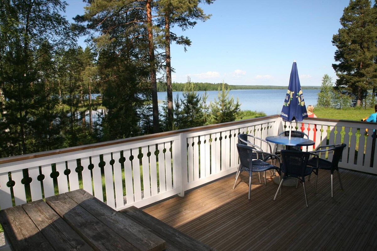 A spacious terrace is visible, featuring a wooden table with four chairs and a large umbrella. The railing is designed with alternating vertical slats, and views of the tranquil lake and surrounding trees can be seen in the background.