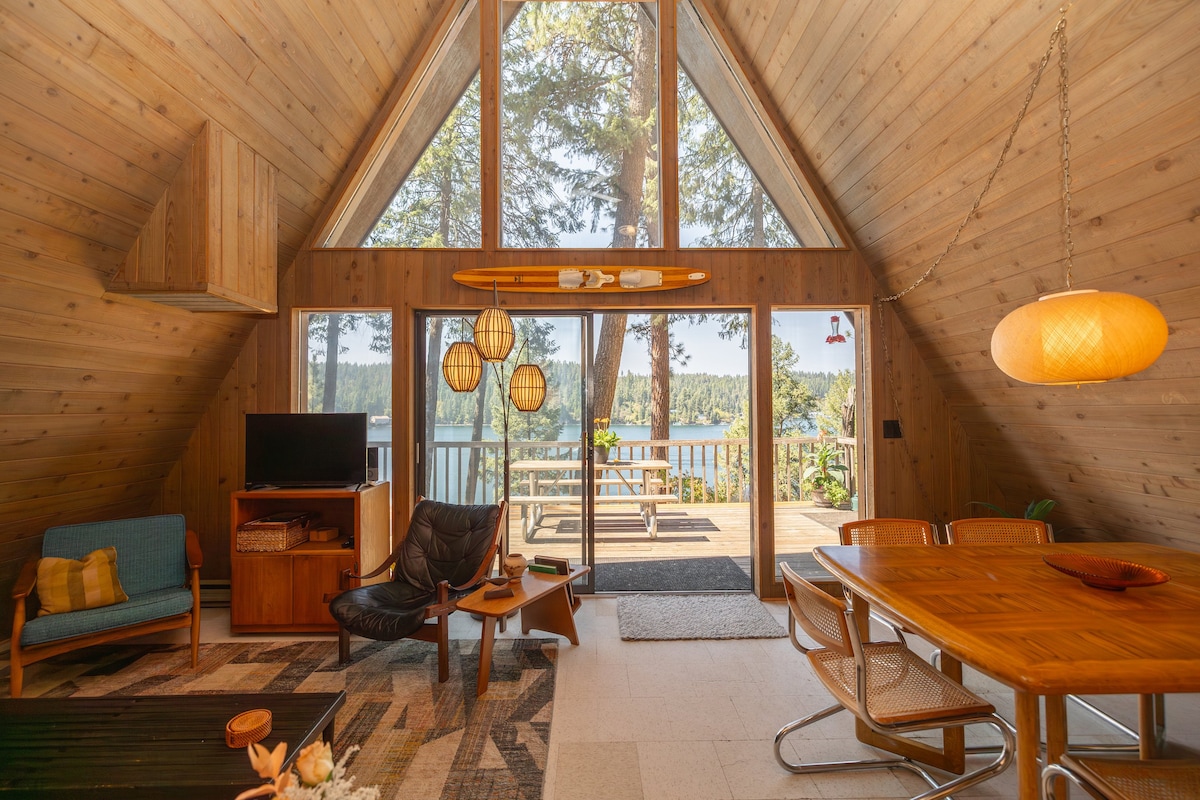 An A-frame cabin interior is showcased, featuring wooden walls and high ceilings. A seating area includes a modern chair and a compact sofa. A dining table with stylish chairs overlooks large windows revealing the lake view and outdoor deck, with a surfboard decoratively displayed.
