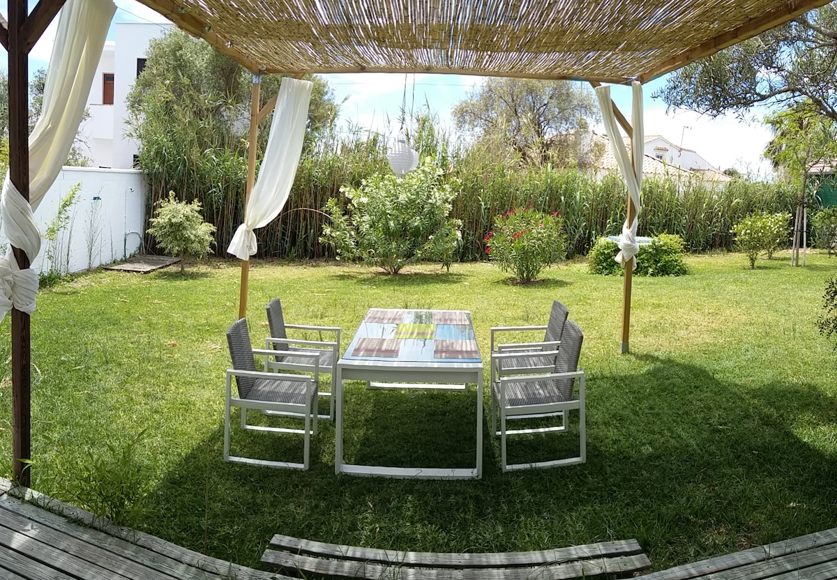 A shaded outdoor dining area is set beneath a woven canopy. A rectangular table is surrounded by four metal chairs, providing a calm space for meals. Lush greenery and flowering plants create a serene backdrop in the fenced-in garden.
