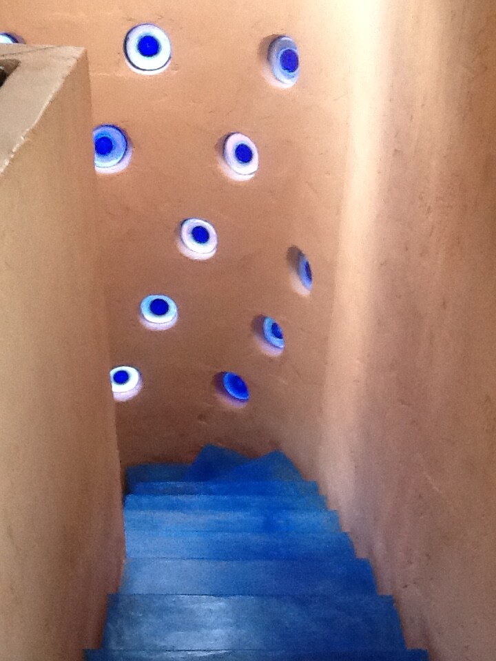 A staircase with blue steps descends into a well-lit area. The walls feature circular, blue-tinted windows that allow natural light to filter through, creating a unique visual texture against the warm-toned plaster.