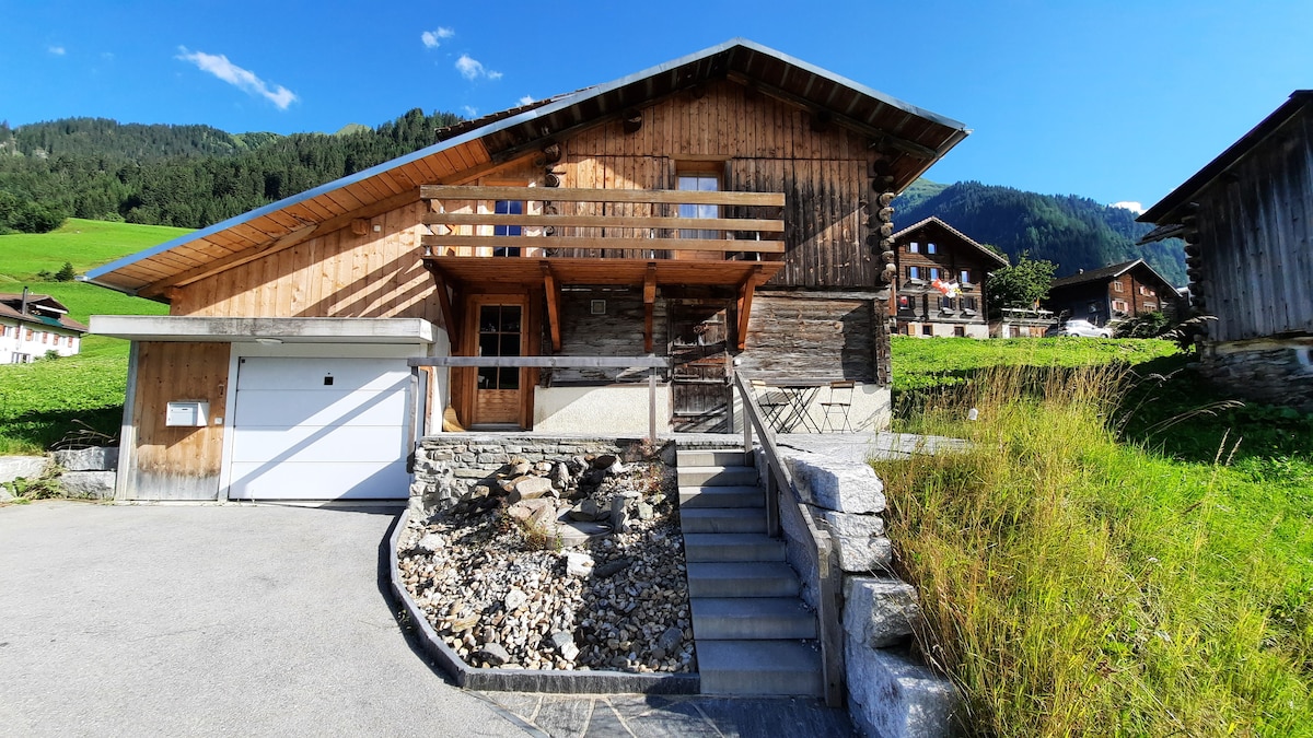 The exterior of a newly renovated chalet features a combination of wooden and stone finishes. A staircase leads to the entrance, flanked by a landscaped area with rocks and grass. A garage is visible, with a scenic backdrop of green hills and distant mountains.