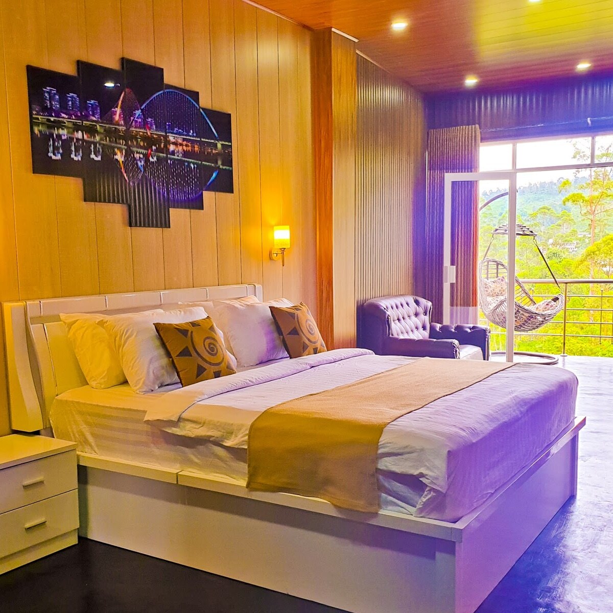 A spacious room features a large bed with decorative pillows, complemented by modern lighting. A cozy seating area is present, furnished with a stylish chair. Floor-to-ceiling windows provide natural light and access to a balcony with views of the surrounding landscape.