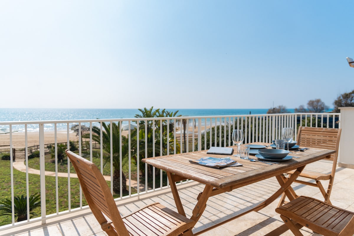 A wooden dining table is set with plates and glasses, positioned on a balcony overlooking the beach and Mediterranean Sea. Sunlight brightens the space, and palm trees can be seen in the distance, complementing the coastal view.