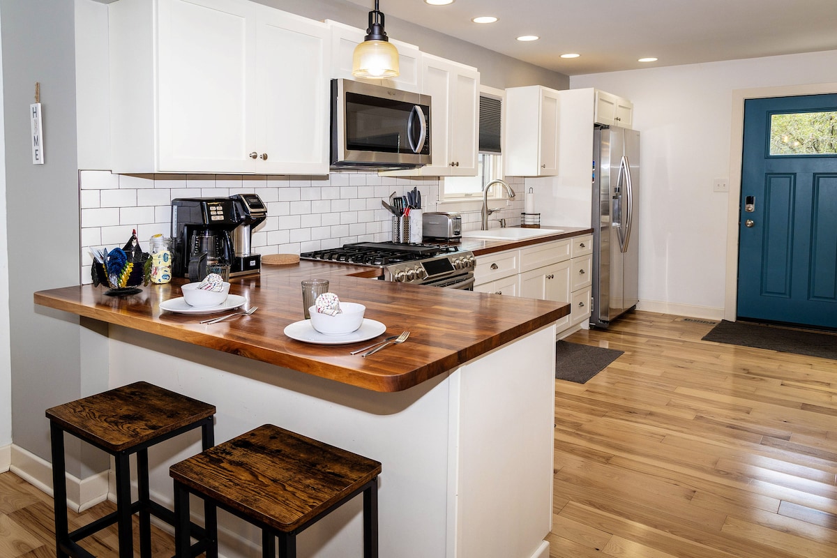 A modern kitchen features light-colored cabinetry, a spacious wooden countertop, and stainless steel appliances. Two black stools are positioned at the counter, and a blue door offers a welcoming entrance. Natural light brightens the space, highlighting the sleek tile backsplash.