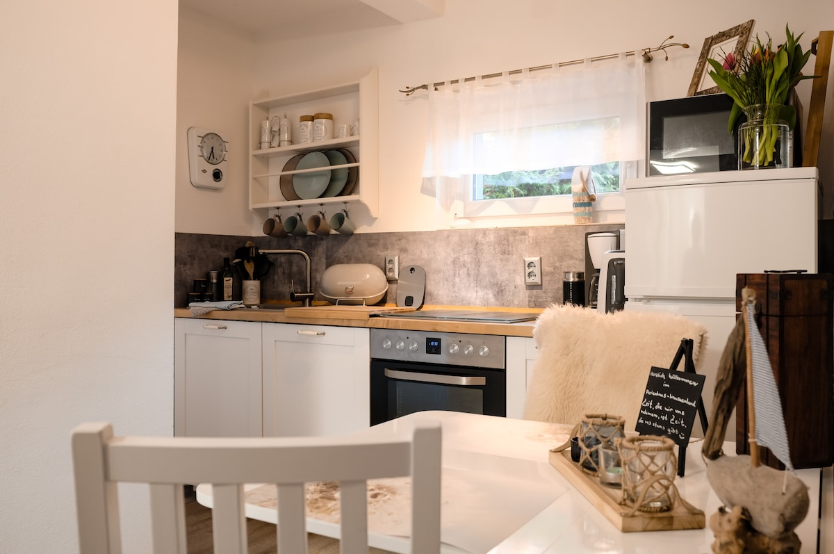 A compact kitchen is equipped with modern appliances, including a stove, oven, and refrigerator. Open shelving displays dining ware and decorative items. A table with light-colored chairs is positioned nearby, complemented by natural light filtering through the window adorned with sheer curtains.