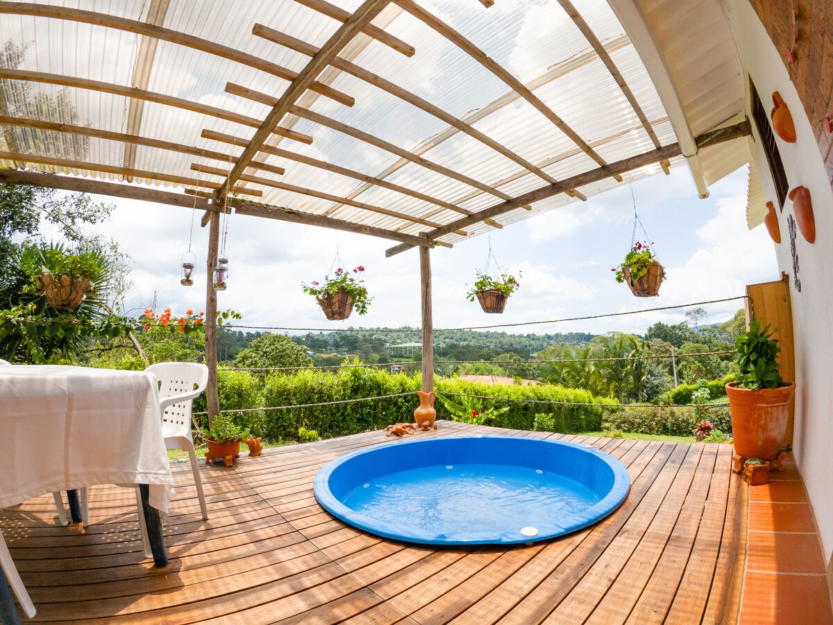An outdoor area features a small blue jacuzzi nestled on wooden decking. Surrounding greenery and vibrant hanging flower pots offer a natural backdrop. A table and chairs are set nearby, inviting guests to enjoy the scenic views of the surrounding mountains and landscape.