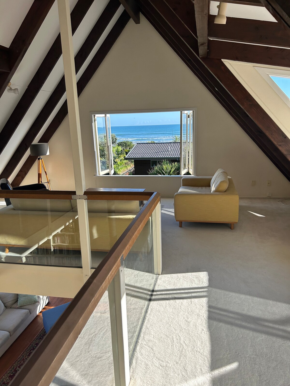 An open loft area is highlighted by wooden beams and large windows offering views of the ocean. A beige sofa is positioned near the window, inviting relaxation. Natural light floods the space, creating a bright and airy atmosphere that connects indoor and outdoor views.