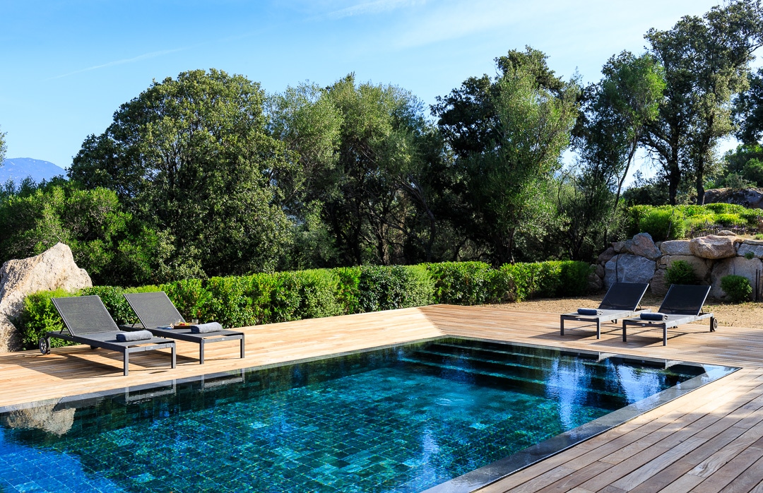 A private salt-water pool is situated on a wooden deck, surrounded by lush greenery and natural rock formations. Sun loungers are positioned neatly beside the pool, inviting relaxation. The clear water reflects the sky, enhancing the serene outdoor environment.