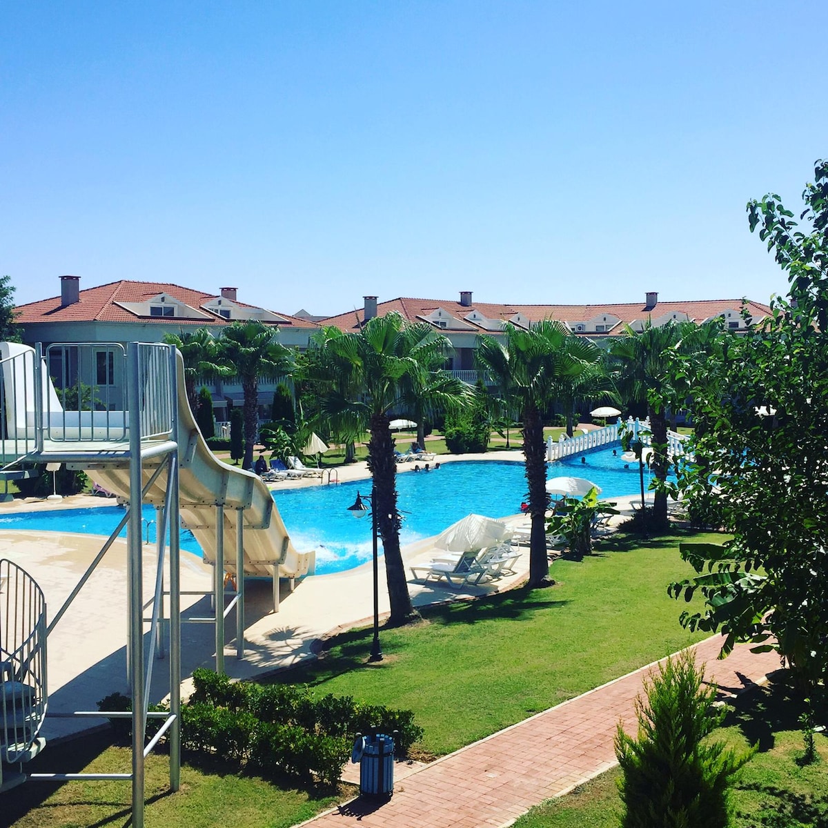 A spacious pool area is surrounded by lush greenery and palm trees. A water slide stands prominently beside the pool, which is divided into various sections for leisure. Chaise lounges are arranged under umbrellas along the pool's edge.