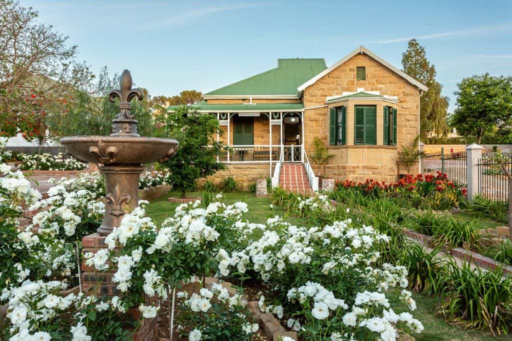 A charming freestanding unit stands surrounded by a well-maintained garden filled with white roses. The facade showcases stonework and green shutters, complemented by a green roof. A decorative fountain adds a serene focus point, while paths lead to the inviting entrance.