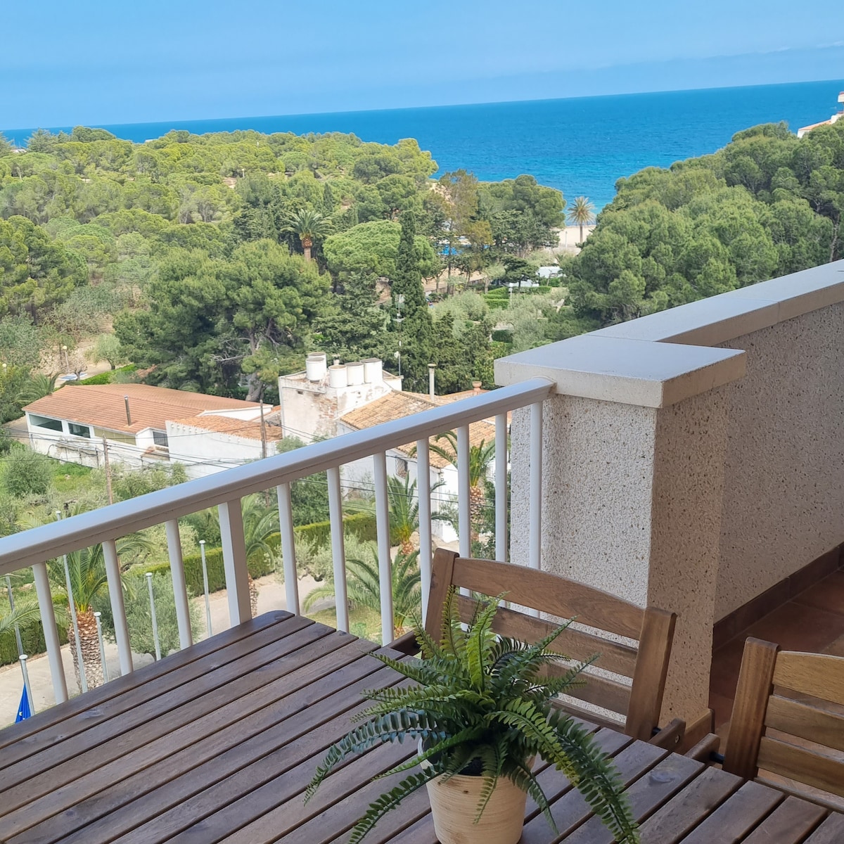 A spacious terrace features a wooden dining table surrounded by matching chairs. A lush green plant sits in the center. In the background, the coastal scenery includes vibrant blue waters meeting a green shoreline, framed by trees and a distant building.