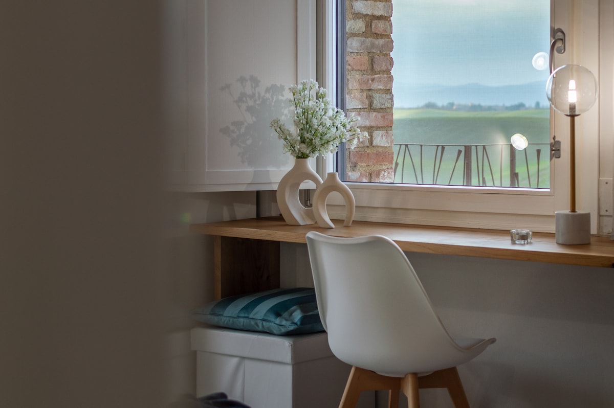 A simple work nook is presented with a white chair and a natural wood desk, positioned near a window. A vase with delicate flowers sits on the desk, with soft light enhancing the serene view of the surrounding landscape.
