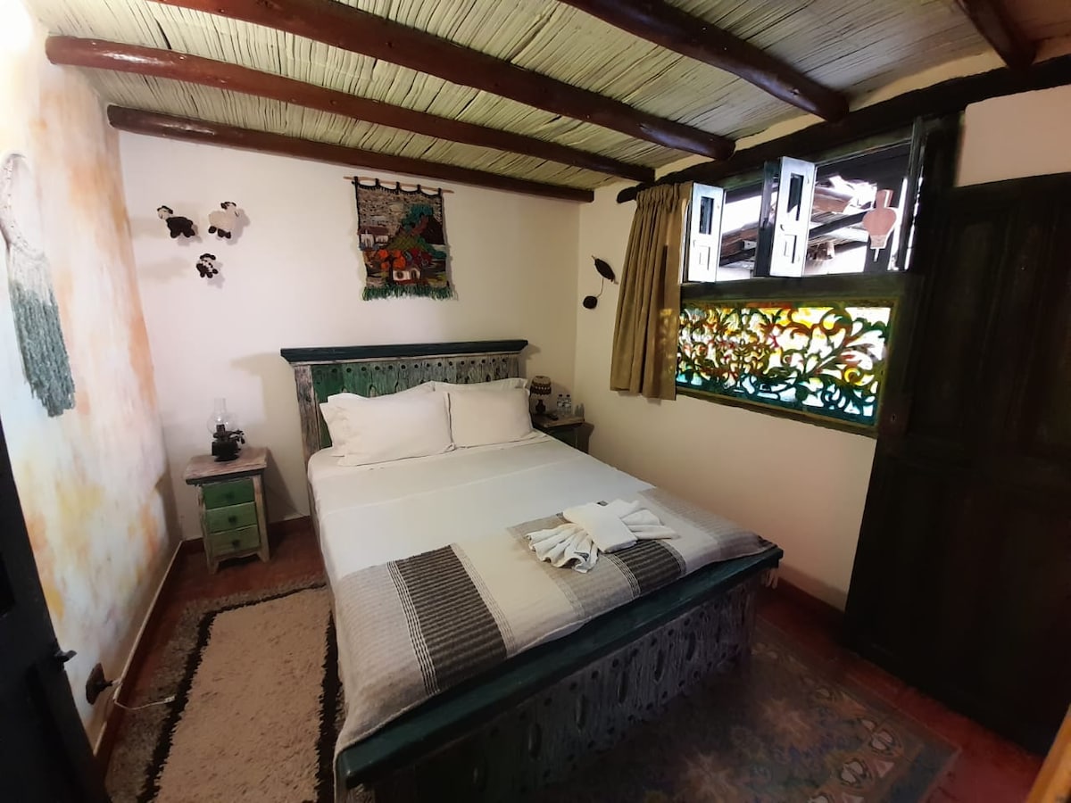 A cozy room features a double bed with crisp linens and a decorative throw blanket. Colorful handcrafted elements adorn the walls, with a vibrant window offering natural light. A wooden closet and a small bedside table add functional charm to the inviting space.