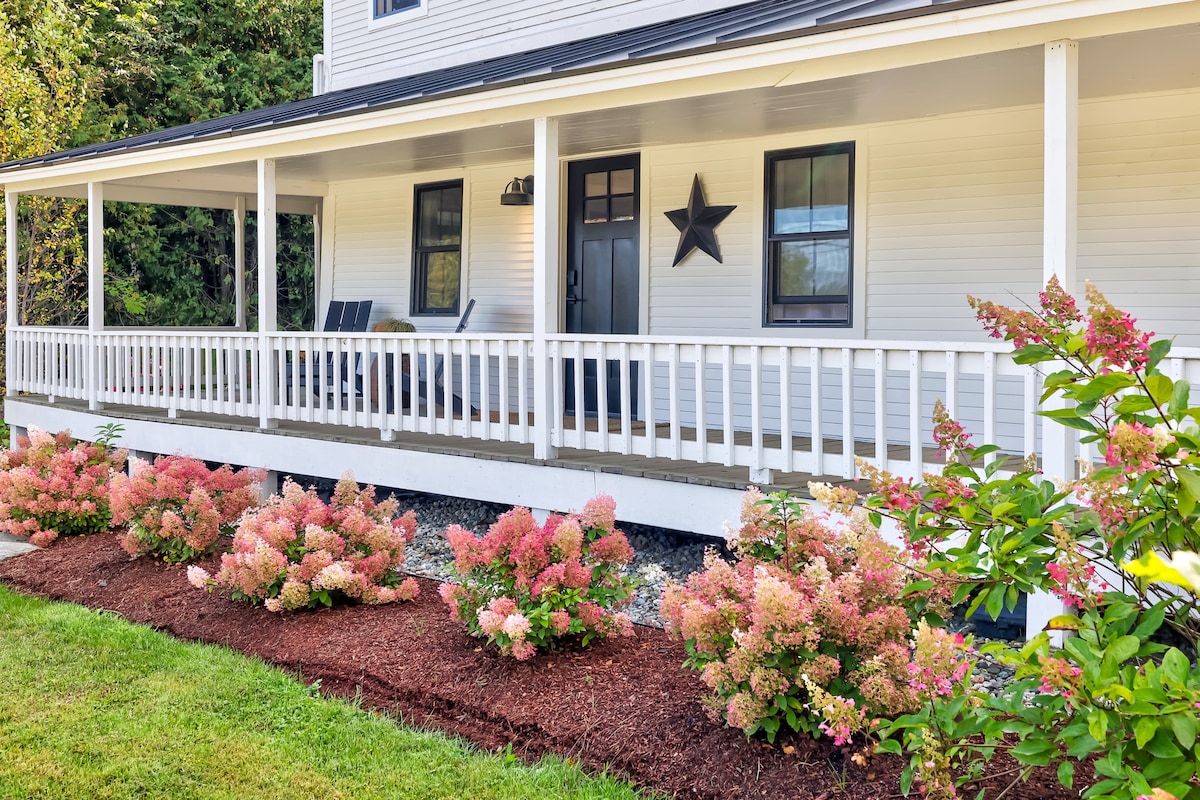 A welcoming front porch features comfortable seating and a decorative star, surrounded by neatly trimmed flower beds with pink hydrangeas. The porch is framed by white railings, offering a charming view of the landscaped yard and nearby greenery.
