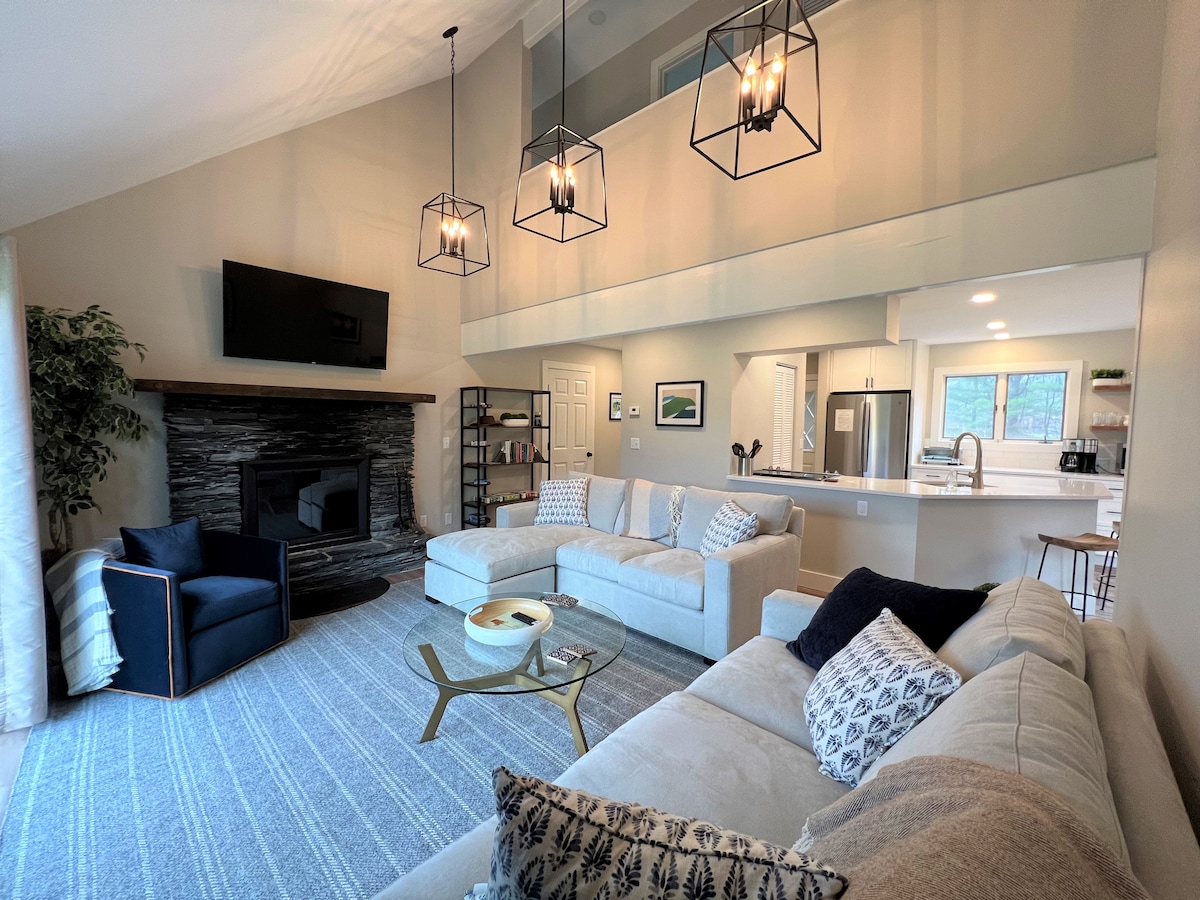 The spacious living area features a comfortable sectional sofa arranged around a glass coffee table, with a cozy blue armchair nearby. A modern wood fireplace provides warmth, complemented by stylish pendant lighting. Large windows allow natural light, showcasing a serene atmosphere with neutral tones.