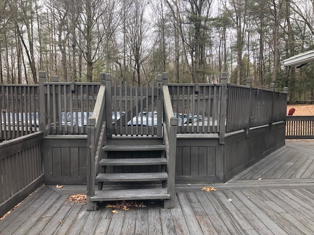 A wooden deck is shown, featuring a set of stairs leading up to a fenced area. The gray wood contrasts with the surrounding trees, indicating a natural and serene setting. Bare branches suggest winter, while leaves lay scattered on the wooden surface.