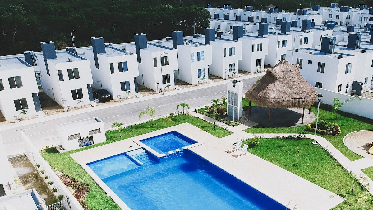 An aerial view of the residential complex is featured, showcasing a rectangular swimming pool surrounded by green grass. A palapa provides shaded seating nearby, with white townhouses visible in the background. The tranquil atmosphere of the area is emphasized by the greenery lining the pathways.