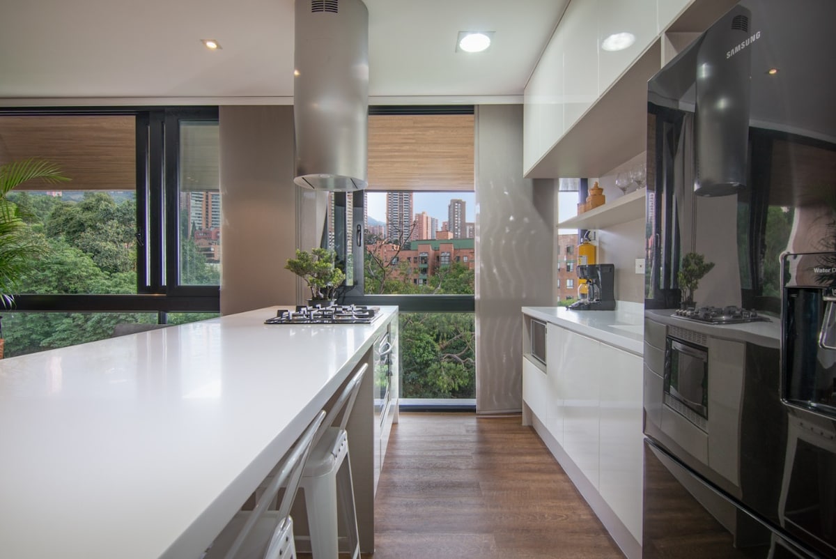 A modern kitchen features sleek cabinetry and a spacious island with barstool seating. Large windows allow natural light to flood the area, providing views of the surrounding greenery and cityscape. A gas stove and sleek black refrigerator are visible, adding to the contemporary design.