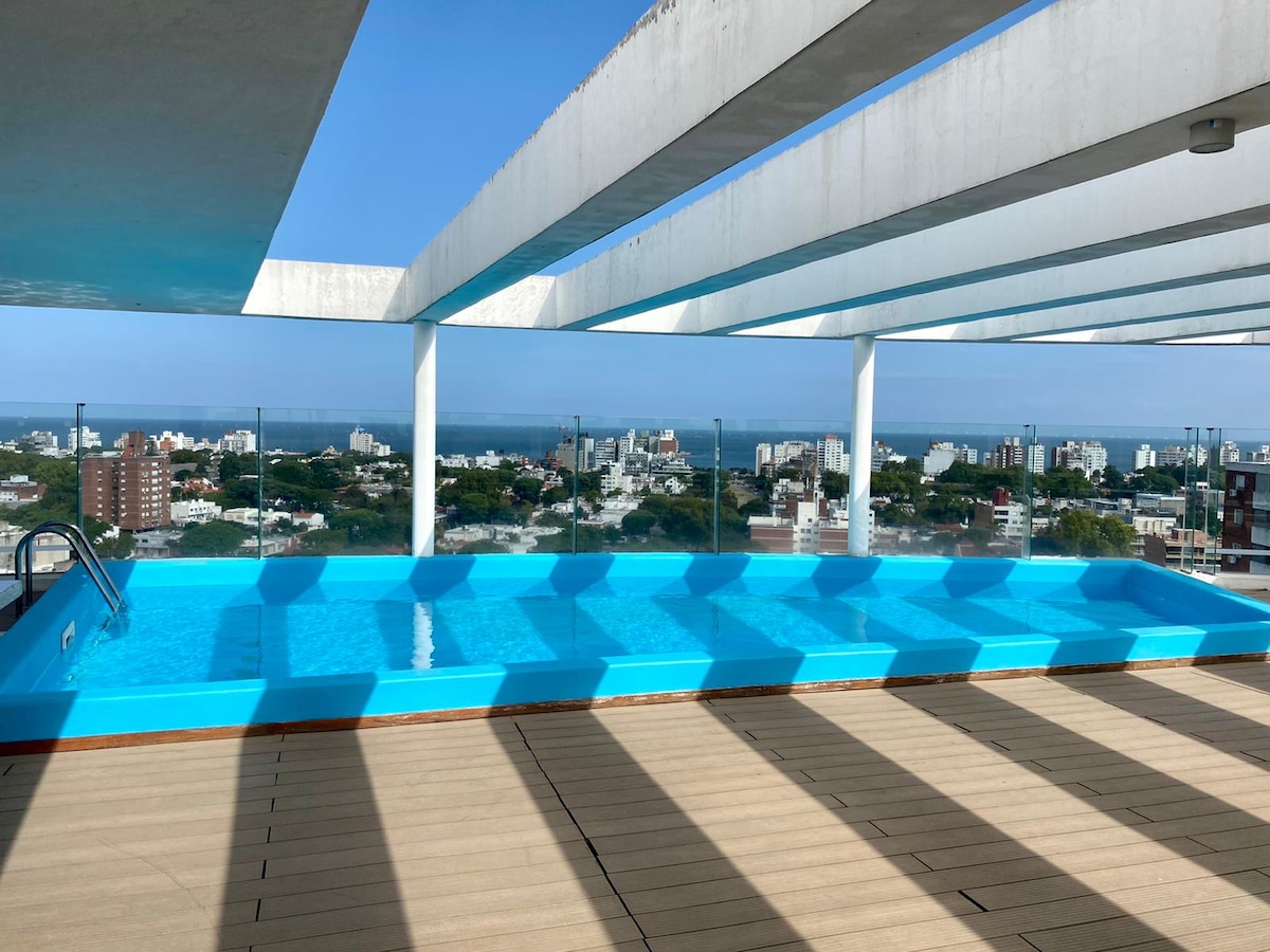 An outdoor pool appears on a rooftop terrace, reflecting sunlight on its turquoise surface. The pool is surrounded by wooden decking and offers expansive views of the city and coastline in the distance, with scattered buildings and greenery visible.