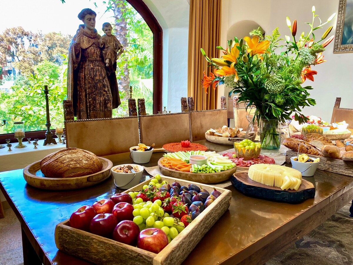 A dining table is set with an array of fresh fruits, cheeses, and breads, complemented by a vase of vibrant flowers. In the background, a stone statue and lush greenery are visible through arched windows, enhancing a warm and inviting dining atmosphere.