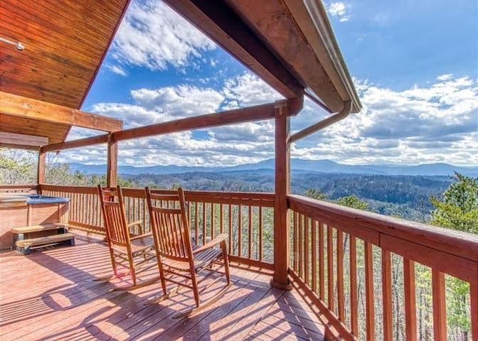 A wooden deck features two rockers overlooking a vast mountainous landscape. The sky is filled with scattered clouds, and rolling hills extend into the distance. A hot tub is positioned nearby, providing an inviting space for relaxation and enjoyment of the serene surroundings.