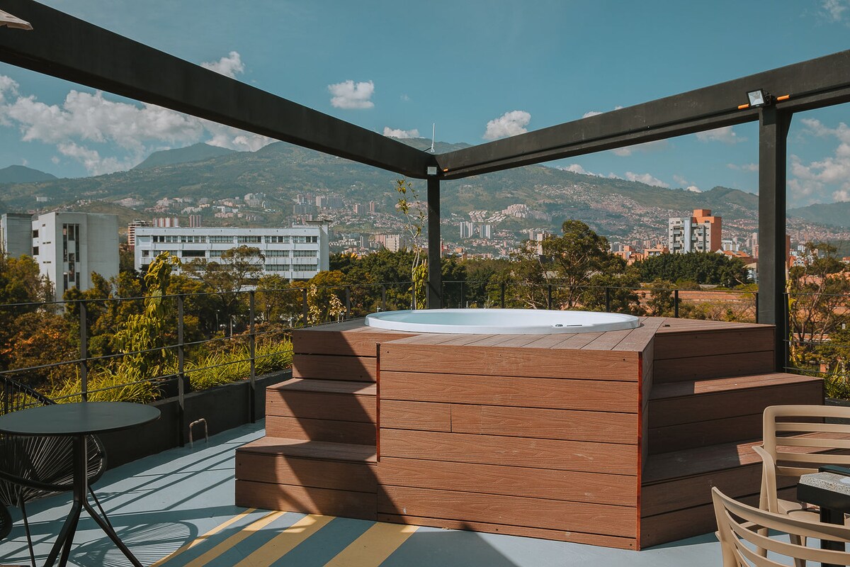 The rooftop social area features a modern jacuzzi, surrounded by wooden decking and a metal frame. Expansive views of the valley and cityscape are visible in the background, under a clear blue sky dotted with fluffy clouds.