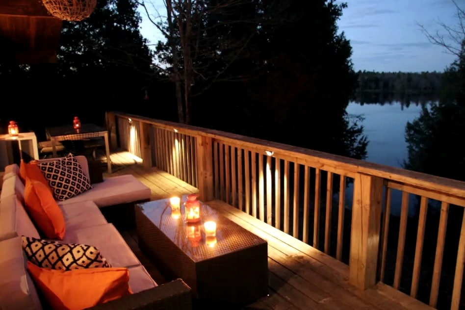 A spacious wrap-around deck is illuminated by soft lighting, creating a cozy ambiance. Comfortable seating with decorative cushions overlooks tranquil lake views, complemented by flickering candles on the table. Surrounding trees provide a sense of privacy, enhancing the serene outdoor atmosphere.