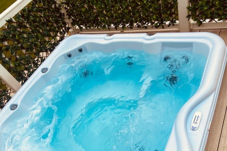 A spacious hot tub is set on the private decking area, featuring bubbling water and comfortably accommodating multiple guests. Surrounded by green foliage, the inviting space offers a serene atmosphere for relaxation.